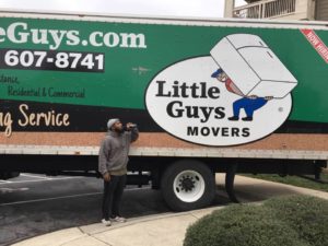 A Little Guy drinks a refreshing beverage in front of a Little Guys Movers moving truck.